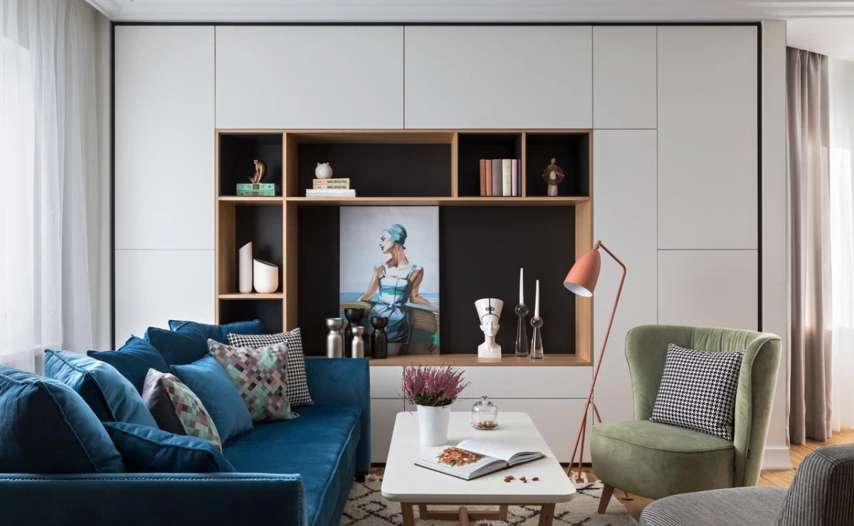 Белые и серые тона и максимально простая отделка в качестве основы позволили удачно расставить яркие акценты. Бордовые стулья, зеленое кресло, синий диван, розовая скамья, необычные картины – вся квартира наполнена цветом, но он не режет глаза. <br><br>

Начиная работу над дизайном интерьера, мы объединили санузел и ванную комнату в одно помещение, а также убрали перегородку в прихожей. Включили в интерьер встроенные шкафы. Это позволило оптимизировать каждый квадратный сантиметр пространства. <br><br>

Система хранения – минималистские встроенные шкафы – почти сливающиеся со стенами, изготовлена по эскизам Натальи Голубович. По эскизам дизайнера изготовлен на заказ и стильный синий диван в гостиной.
<br><br>
Зона гостиной построена таким образом, чтобы заказчица могла расслабиться и отдохнуть, вести долгие разговоры по душам и мечтать, а не банально смотреть телевизора. Самое интересное здесь – отсутствие верхнего света, только торшер и настольные лампы. <br><br>

Мы задумали максимально чистый, незагроможденный проект, без обременения большим количеством элементов. Наш проект дизайна интерьера однокомнатной квартиры в Минске доказывает: даже небольшое жизненное пространство можно сделать функциональным, удобным, современным!
